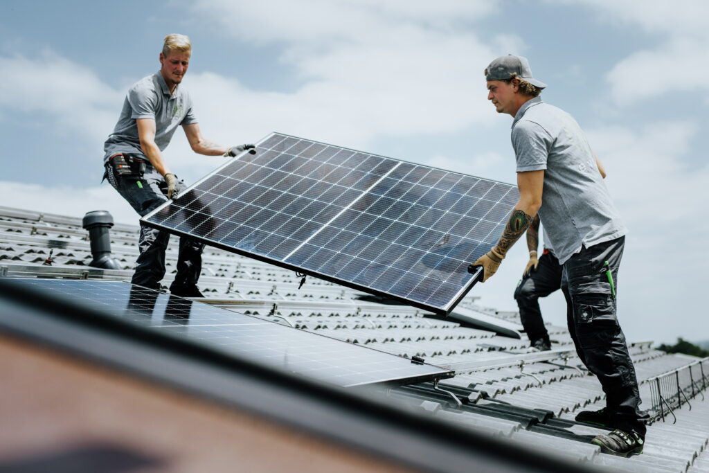 Zeigt 2 Personen, welche ein PV-Modul auf dem Dach tragen. 