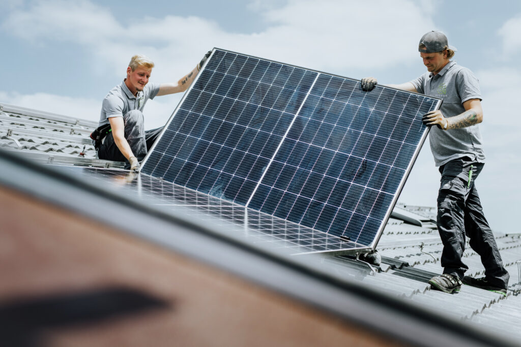 Zeigt zwei Personen auf einem Hausdach welche ein PV-Modul halten. 